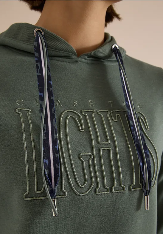 Grüner Hoodie mit geprägtem ‚CHASE LIGHTS‘-Schriftzug und gemusterten Kordeln, nur Oberkörper sichtbar.