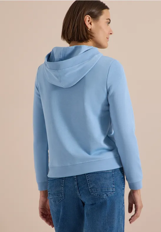 Eine Frau von hinten in hellblauem Hoodie und Jeans, Blick nach rechts.