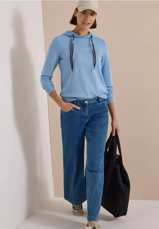 Frau in hellblauem Hoodie, weitem Jeans, weißer Cap und Tasche, stehend vor beigem Hintergrund