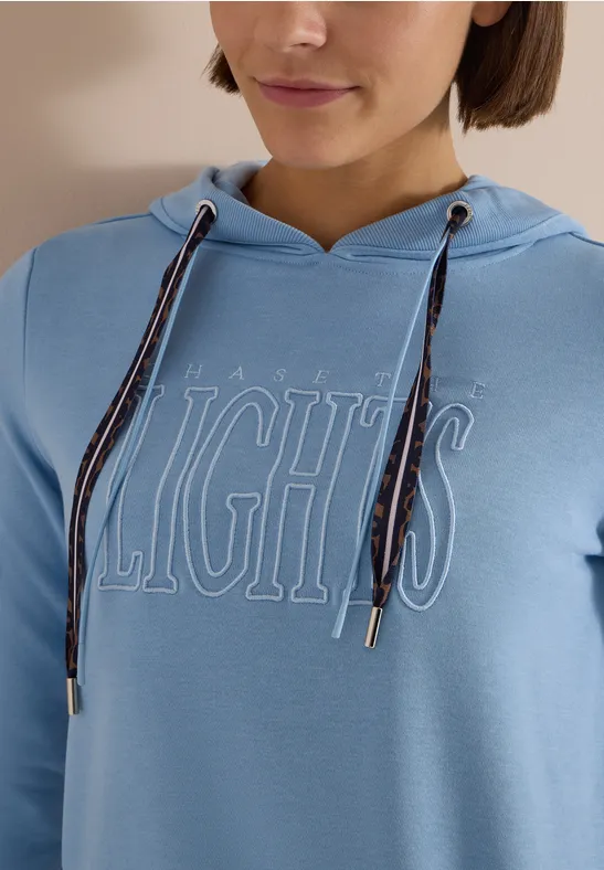 Person trägt hellblauen Kapuzenpulli mit weiß umrissenem 'LIGHTS'-Schriftzug und gestreiften Kordeln.