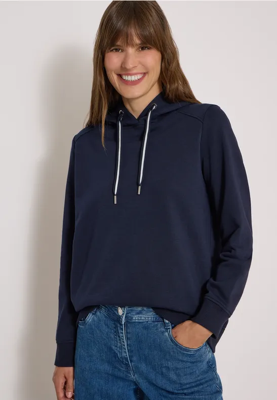 Eine Frau mit langen braunen Haaren trägt lässigen dunkelblauen Kapuzenpullover und Jeans, vor hellem Hintergrund.