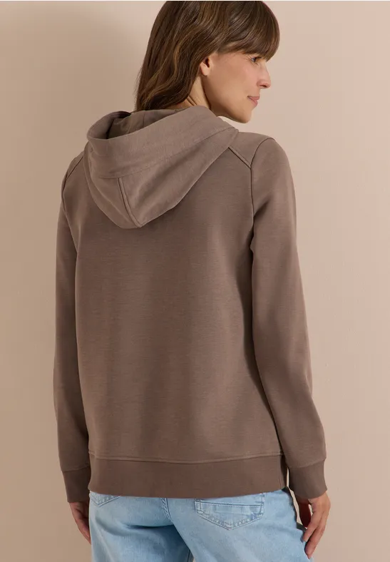 Frau von hinten in braunem Kapuzenpullover und blauen Jeans, leicht seitlich gedreht.