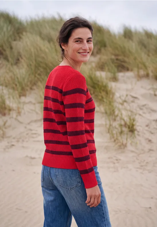 Een glimlachende vrouw in een rood-zwart gestreepte trui staat op het strand voor duinen.