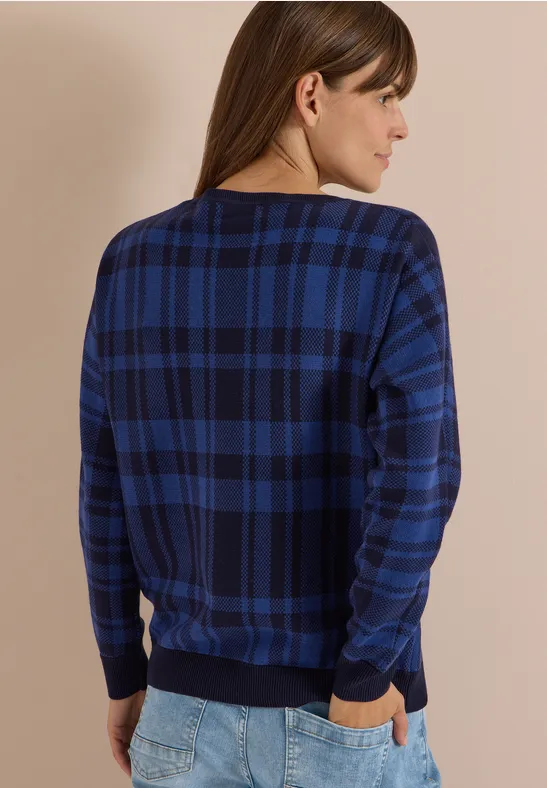 Frau von hinten in dunkelblau kariertem Pullover und Jeans, Hände in den Taschen.
