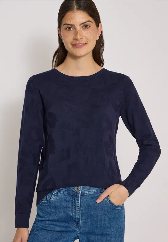 Junge Frau mit dunklen Haaren trägt ein dunkelblaues Langarmshirt und Jeans, lächelnd vor weißem Hintergrund.