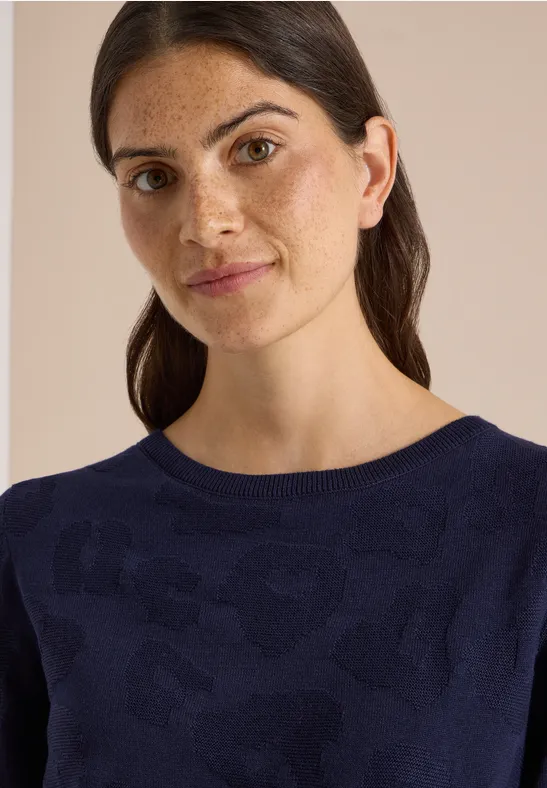 Porträt einer Frau mit dunklen Haaren und Sommersprossen in marineblauem Pullover vor beigem Hintergrund.