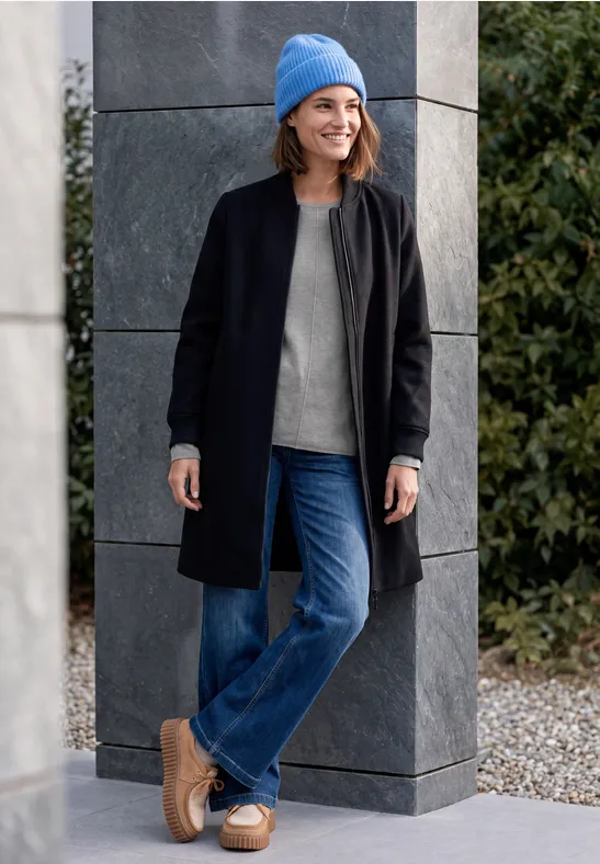 Eine Frau in schwarzem Mantel, Jeans, grauem Pullover und blauer Mütze lehnt lächelnd an einer Säule im Freien.