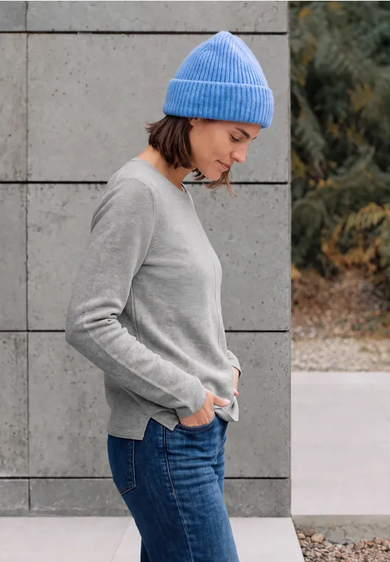 Frau mit blauem Beanie und grauem Pullover steht seitlich vor einer Betonwand.