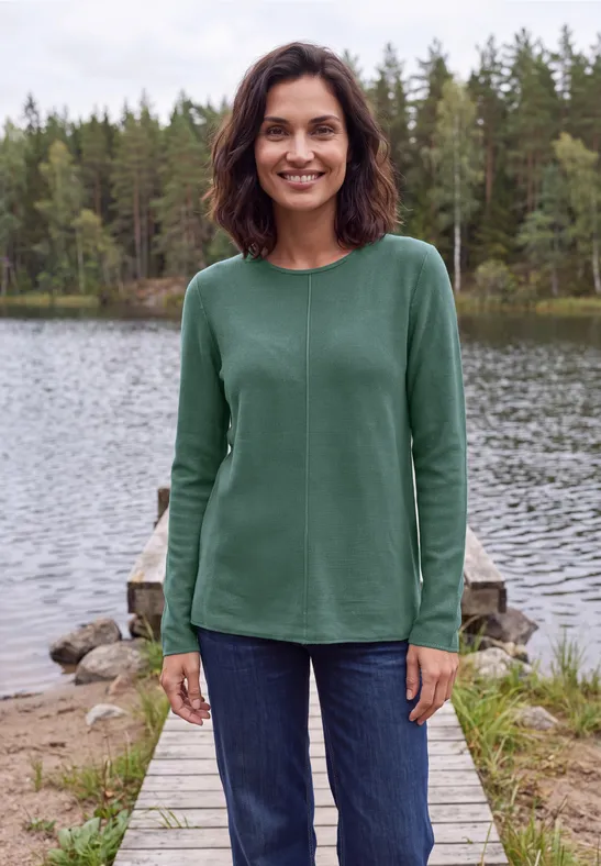 Eine lächelnde Frau in grünem Pullover und Jeans steht auf einem Steg am See, umgeben von Wäldern.