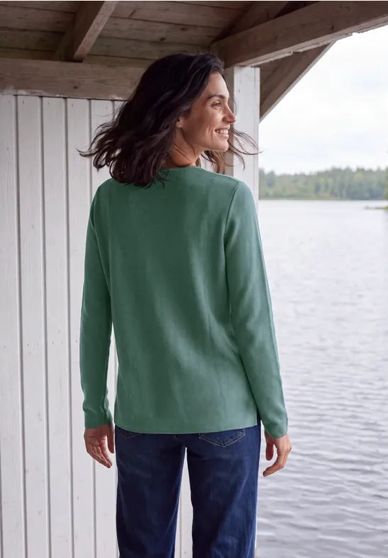 Frau im grünen Pullover steht auf einer überdachten Terrasse und blickt auf einen See.