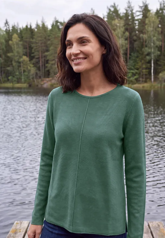 Eine lächelnde Frau trägt einen grünen Pullover am Ufer eines Sees mit Wald im Hintergrund.