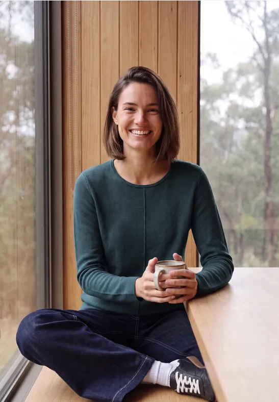 Een vrouw zit in lotushouding op een bank bij het raam, houdt een mok vast en lacht ontspannen.