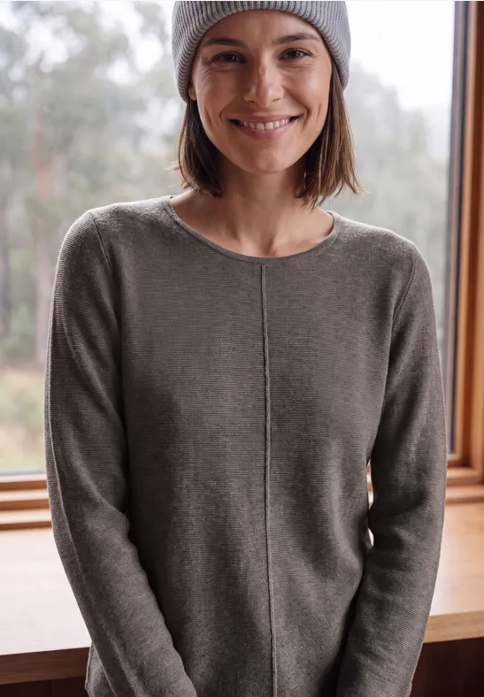 Eine lächelnde Frau in einem grauen Pullover und Mütze steht vor einem Fenster mit Waldblick.