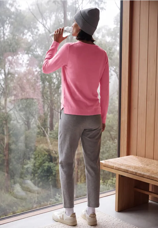 Eine Person in rosa Pullover, grauer Mütze und Hose steht an einem Fenster und trinkt aus einer Tasse.