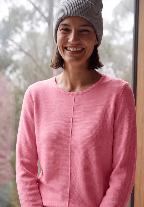 Junge Frau in rosa Pullover und grauer Mütze, die vor einem Fenster lächelt