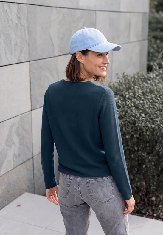 Frau in Jeans, Pullover und hellblauer Baseballkappe lehnt an Mauer und blickt zur Seite