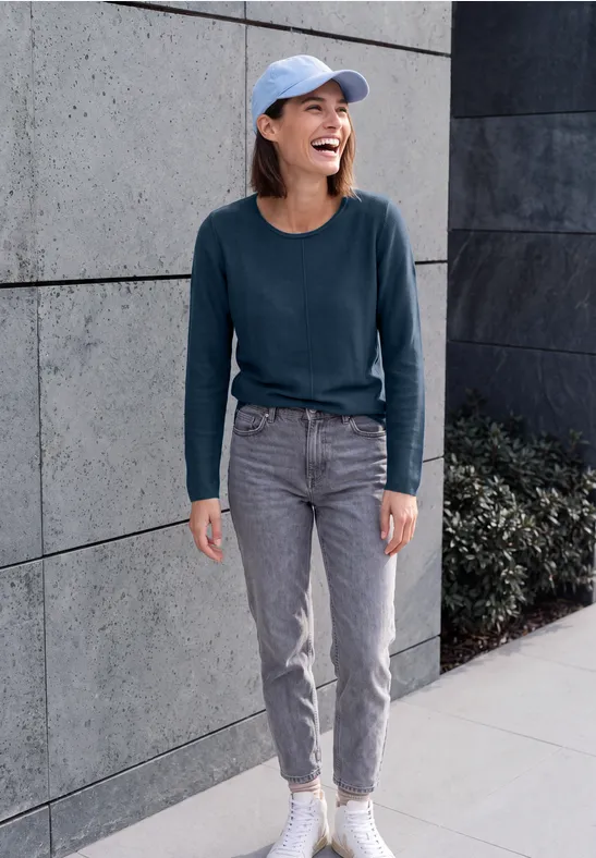 Lächelnde Frau in Jeans, Pullover und Baseballkappe vor grauer Mauer.