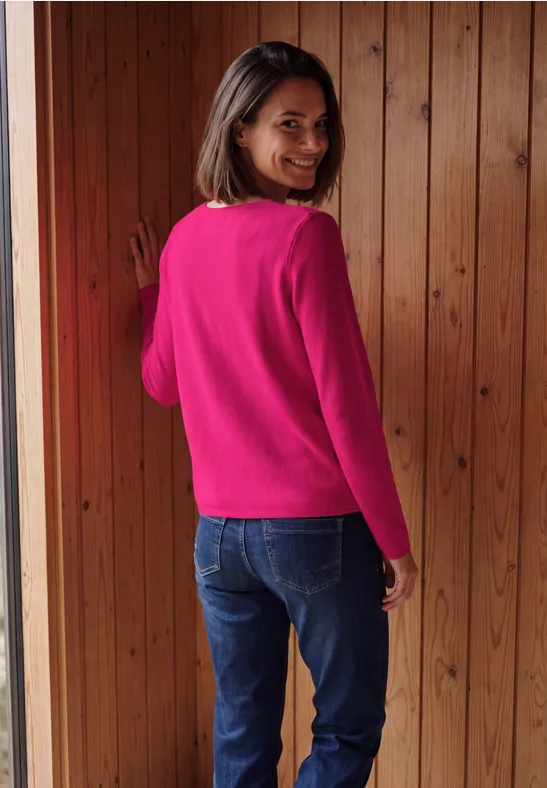 Eine Frau in pinkem Pullover und Jeans steht vor einer Holzwand und lächelt über die Schulter.