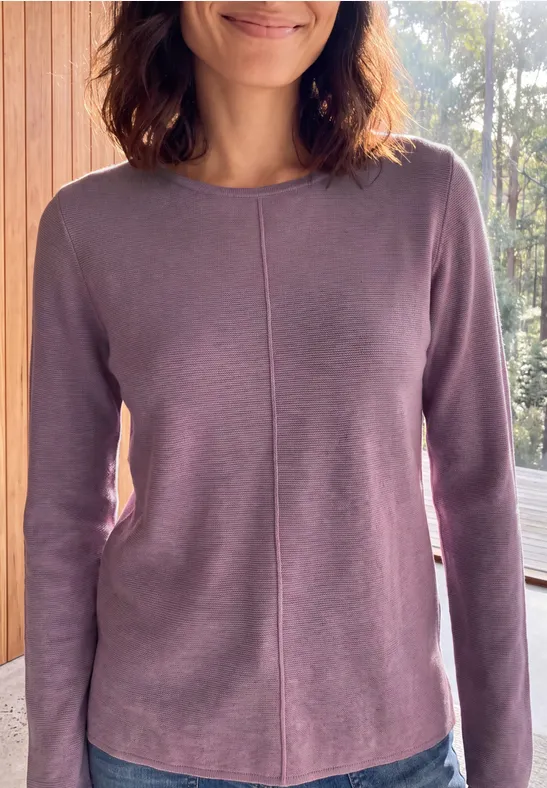Femme en pull violet debout dans une pièce lumineuse devant une fenêtre.