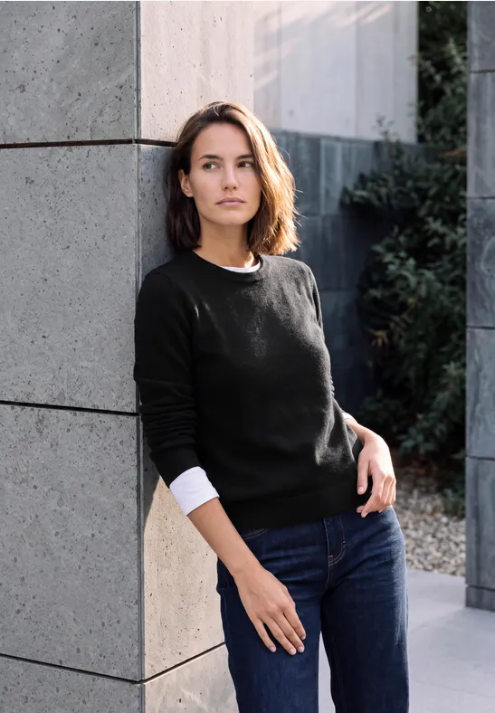 Une femme appuyée contre un mur en pierre clair, vêtue d’un pull noir et d’un jean, posant à l’extérieur près de plantes.