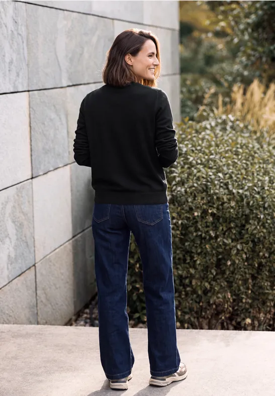 Femme en pull noir et jean bleu, vue de dos, debout devant un mur et des buissons.