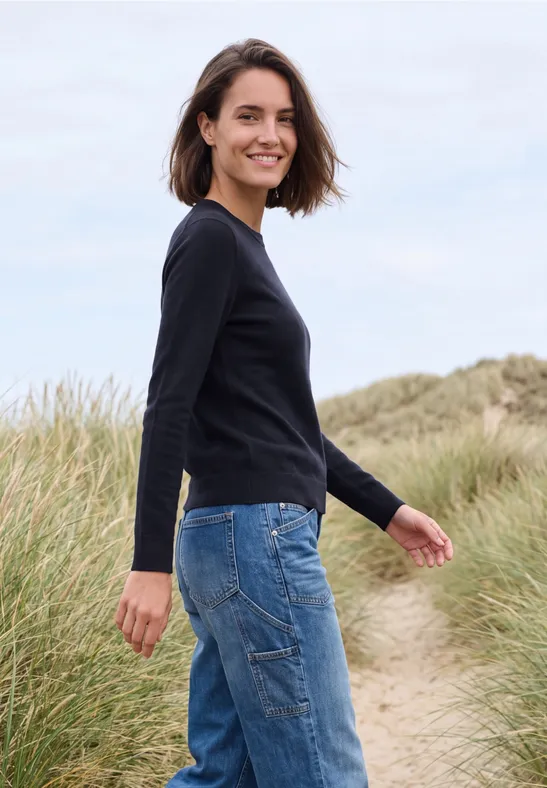 Eine lächelnde Frau in schwarzem Pullover und Jeans spaziert durch Grasdünen am Strand.