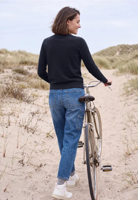 Frau schiebt Fahrrad in Jeans, schwarzem Pullover und weißen Sneakers über Dünenpfad am Strand