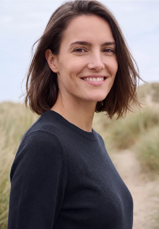 Eine lächelnde Frau mit braunen Haaren in schwarzem Pullover vor Dünenkulisse.
