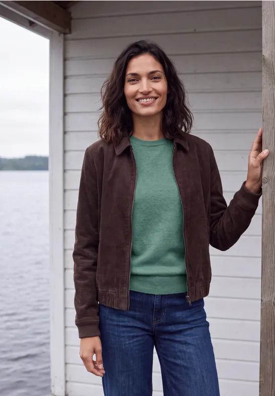 Lächelnde Frau in grünem Pullover, brauner Jacke und Jeans steht an einem Holzhaus am Wasser.