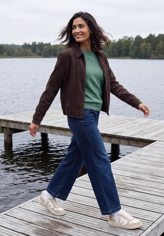 Frau in grünem Pullover und brauner Jacke spaziert lachend auf einem Steg am See.