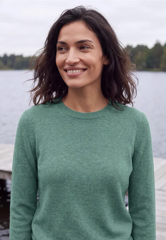 Eine lächelnde Frau im grünen Pullover vor einer See-Kulisse.