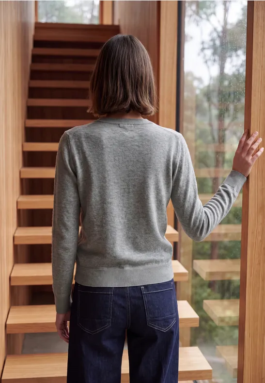Eine Person in grauem Pullover und Jeans steht mit dem Rücken zur Kamera vor einer Holztreppe.