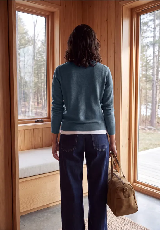 Une femme en pull et jean, sac à la main, se tient dans une cabane en bois et regarde par la fenêtre.