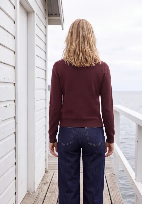 Eine Frau steht von hinten auf einer Strandveranda, trägt einen bordeauxroten Pullover und blaue Jeans.