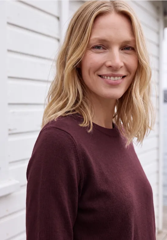Eine lächelnde blonde Frau in einem weinroten Pullover vor einer weißen Wand.