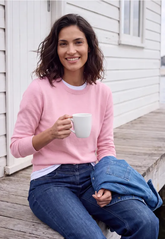 Eine lächelnde Frau in rosa Pullover und Jeans sitzt auf einer Holzstufe, hält eine weiße Tasse.