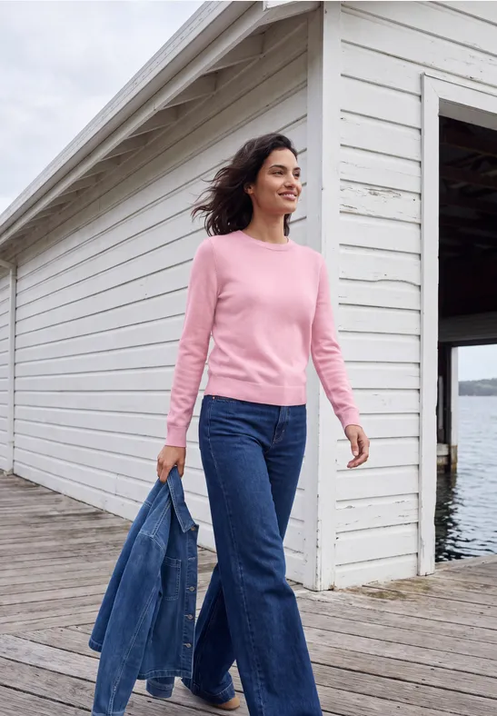 Frau in rosa Pullover und Jeans spaziert mit Jeansjacke an einem Steg vor Bootshaus.
