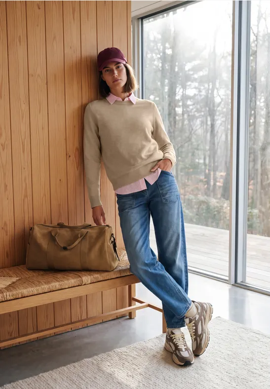 Eine Frau in Pullover, Jeans und Cap lehnt entspannt an einer Holzwand neben einer Reisetasche.