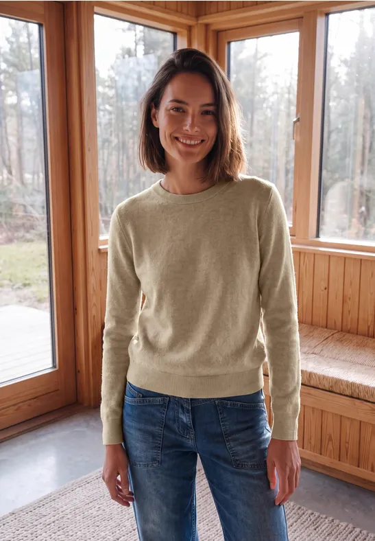 Eine lächelnde Frau in beigem Pullover und Jeans steht in einem holzverkleideten Raum vor großen Fenstern