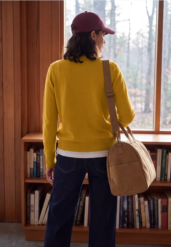 Person mit roter Kappe und senfgelbem Pullover steht vor Fenster und Bücherregal, trägt Tasche.