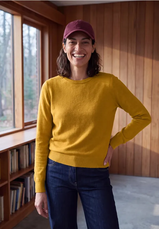 Frau in gelbem Pullover, blauen Jeans und rotem Cap, lächelnd neben Fenster in Holzinterieur.