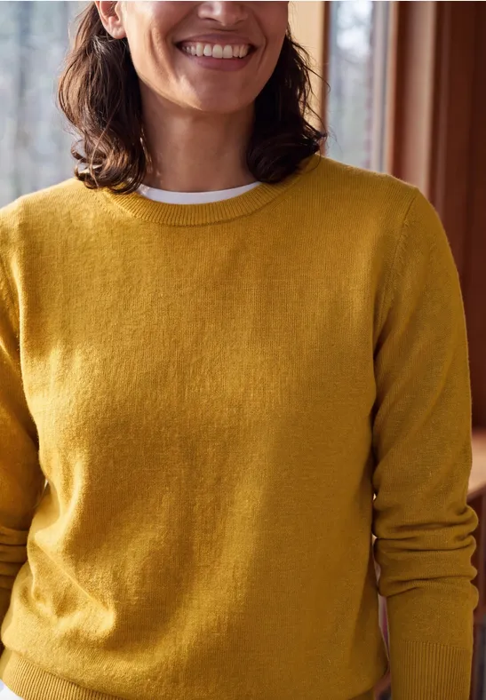 Eine lächelnde Frau trägt einen gelben Pullover in einem gemütlichen Innenraum.
