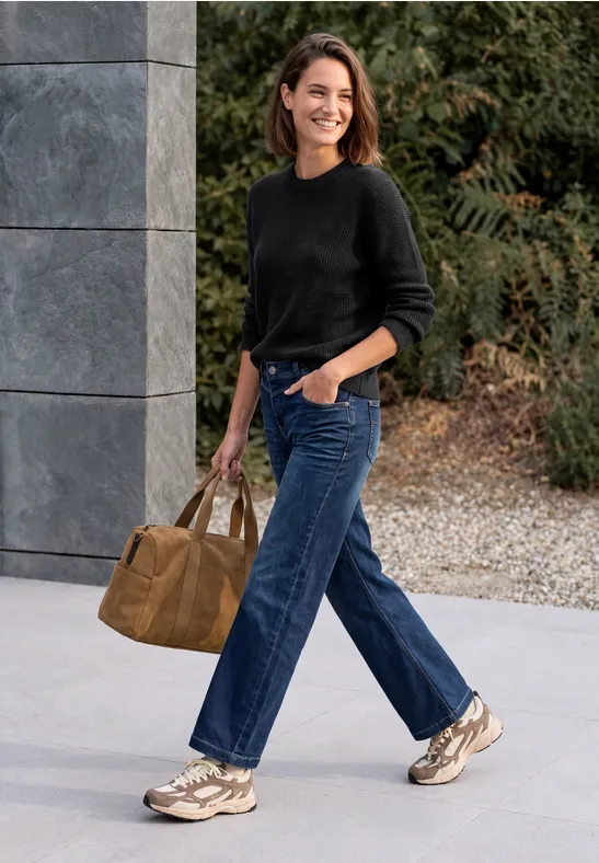 Une femme souriante en pull noir, jean bleu et baskets marche en plein air avec un sac marron.