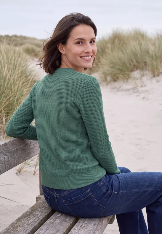 Frau sitzt auf einer Strandbank vor Dünengras, blickt zurück, trägt grünen Pullover und Jeans.