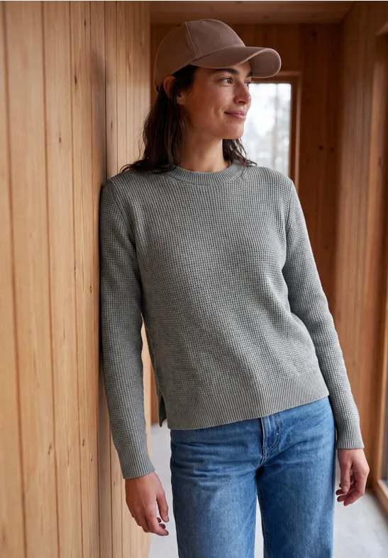 Eine Frau lehnt in grauem Pullover, blauer Jeans und Cap an einer Holzverkleidung und lächelt.