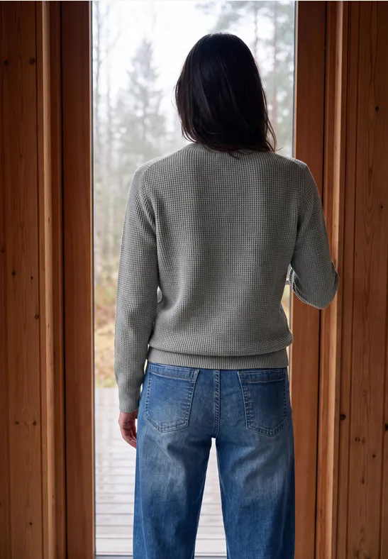 Person mit grauem Pullover und blauen Jeans blickt von einer Holztür aus in den herbstlichen Wald.