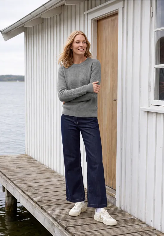 Frau in grauem Pullover und weiten Jeans steht auf einem Steg vor einem weißen Holzhaus am Wasser.