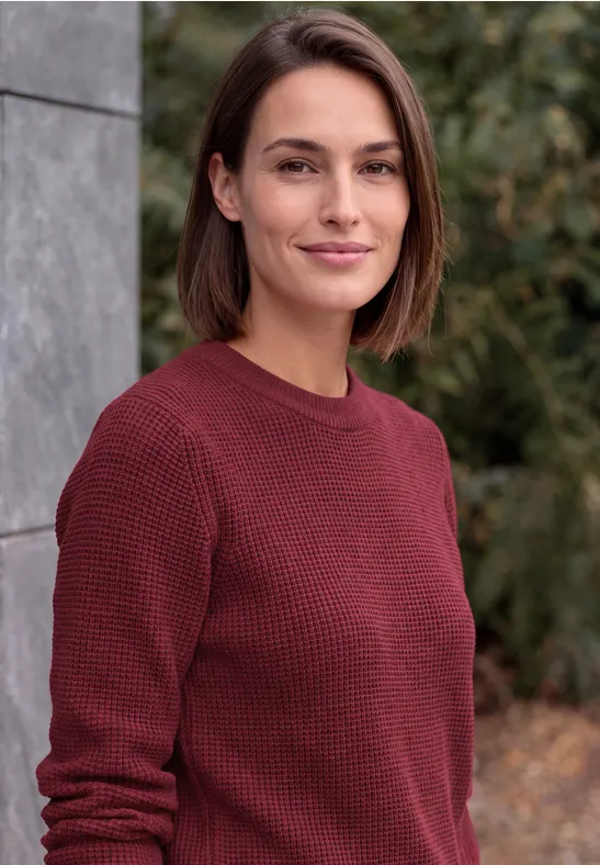 Eine Frau mit kinnlangen braunen Haaren und bordeauxrotem Pullover im Freien.