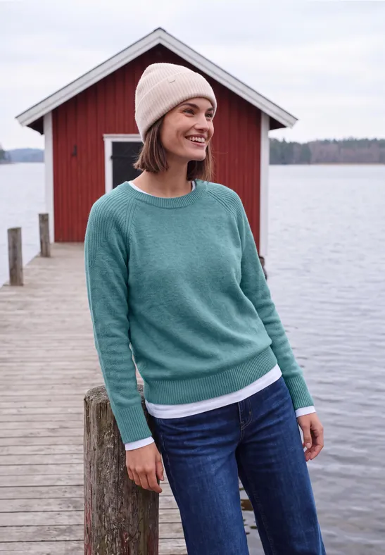 Lächelnde Frau im grünen Pullover, Jeans und Mütze auf einem Steg vor rotem Bootshaus am See.