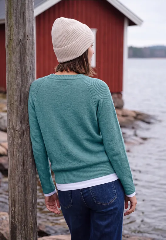 Person mit grüner Strickjacke und Beanie steht rückwärts am Wasser vor einem roten Gebäude.